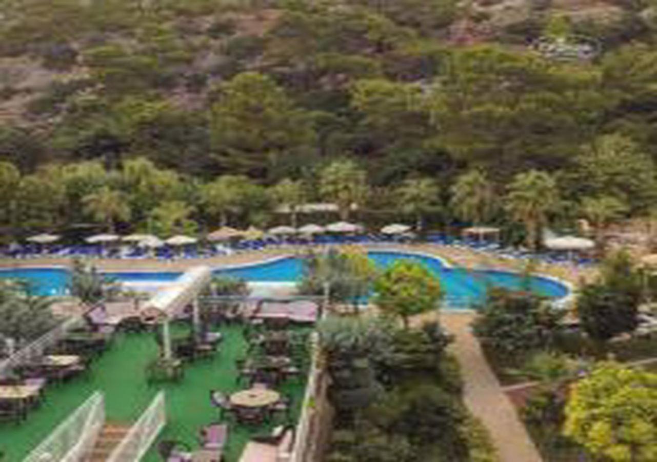 Miramor Hotel & Spa Antalya Luaran gambar The photo shows a scenic view of a resort area featuring a large swimming pool surrounded by lush greenery and trees. Sun loungers are arranged around the pool, and there are tables visible in a dining area. The pool has a curved shape and is complem